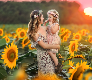 Mother holding toddler daughter in a sunflower field at golden sunset, both wearing floral crowns.