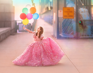A girl in a pink ball gown holds colorful balloons on a sunlit urban sidewalk.