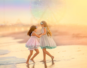 Two young girls in pastel dresses dancing on a beach at sunset with a rainbow in the background.