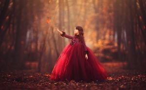 A girl in a red ball gown reaches out to a falling leaf in an autumn forest.