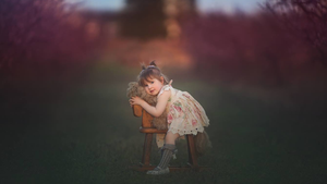 A little girl in a floral dress leans over a wooden chair in a dreamy blossoming garden.
