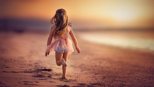A carefree little girl in a pink dress runs barefoot along a sandy beach at sunset.