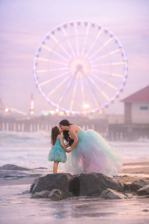 A mother in a rainbow tulle gown kisses her daughter on rocks near a lit Ferris wheel.