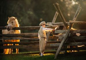 A young boy in a hat reaches out to a palomino horse over a rustic wooden fence at sunset.