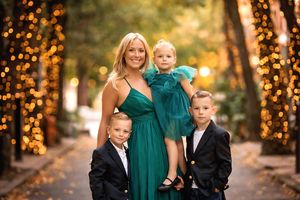 A blonde mother in a green gown poses with her three children amid warm bokeh lights outdoors.