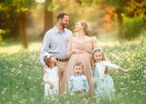 A pregnant mother and father pose with three young children in a sunny flower field.