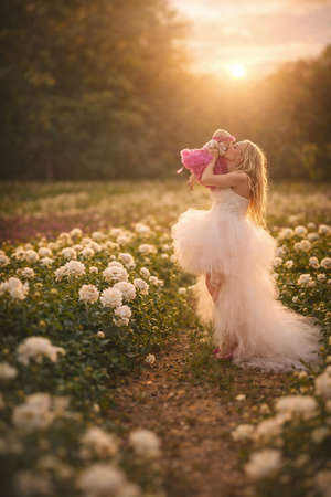 A woman in a white gown kisses a baby in a pink flower field at sunset.