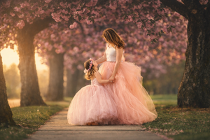 A mother in a pink tulle gown embraces her daughter under blooming cherry blossom trees at sunset.