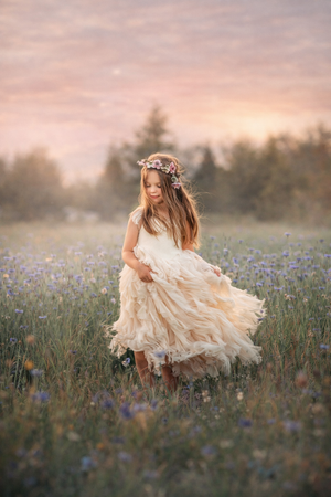 A young girl in a cream lace dress and floral crown twirls in a meadow at sunset.