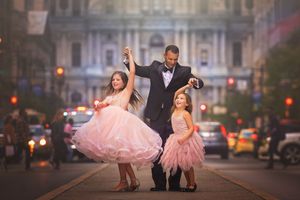 A father dances with his two daughters in pink tulle dresses on a city street.