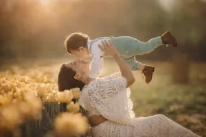 A mother in a white lace dress lifts her laughing toddler in a field of yellow tulips.