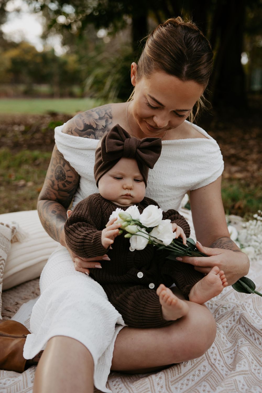 Tattooed mother sitting outdoors holding baby in brown knit outfit with bow headband and white flowers.