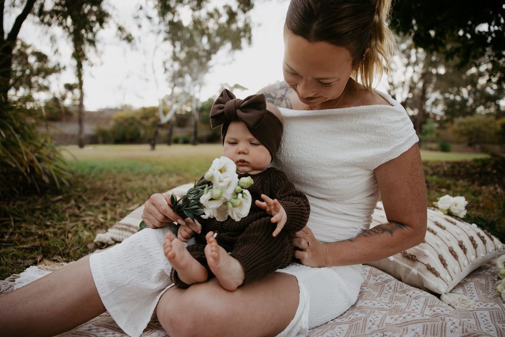 Mother in white dress holds baby with white flowers on a picnic blanket outdoors.