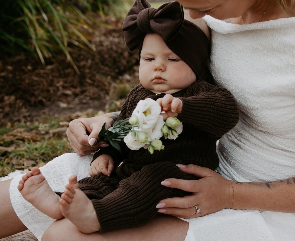 Baby in brown knit outfit with bow headband holds white flowers while sitting on mother's lap outdoors.