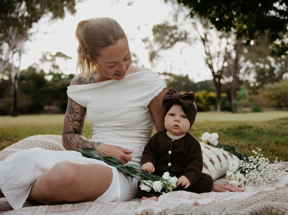 Mother and baby sitting on blanket in park, baby wearing dark outfit with bow headband holding white flowers.