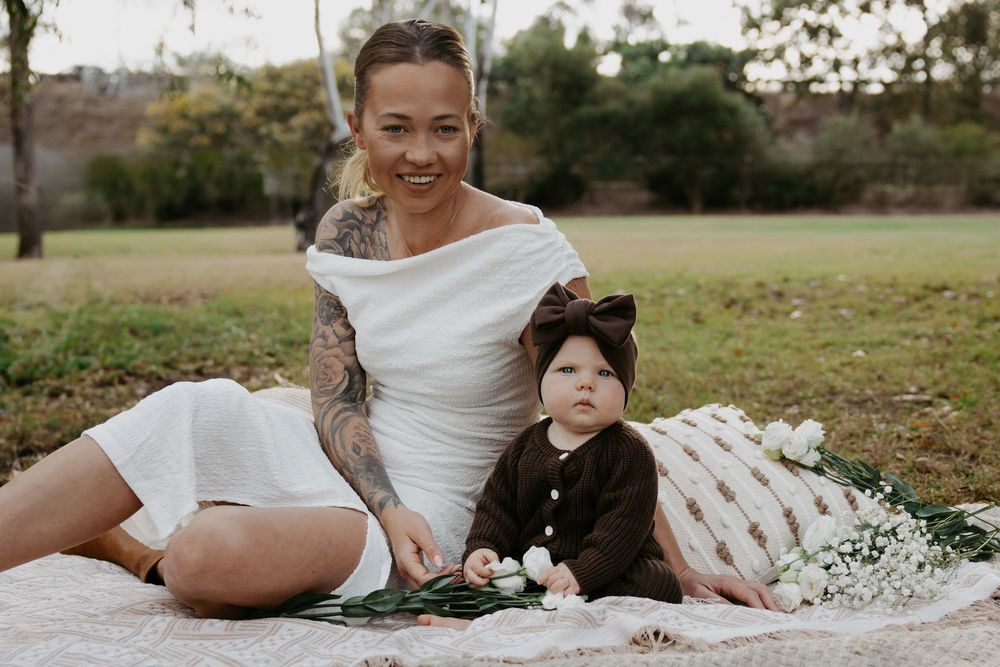 A tattooed woman in a white dress sits on a blanket outdoors with a baby in a brown outfit and bow headband.