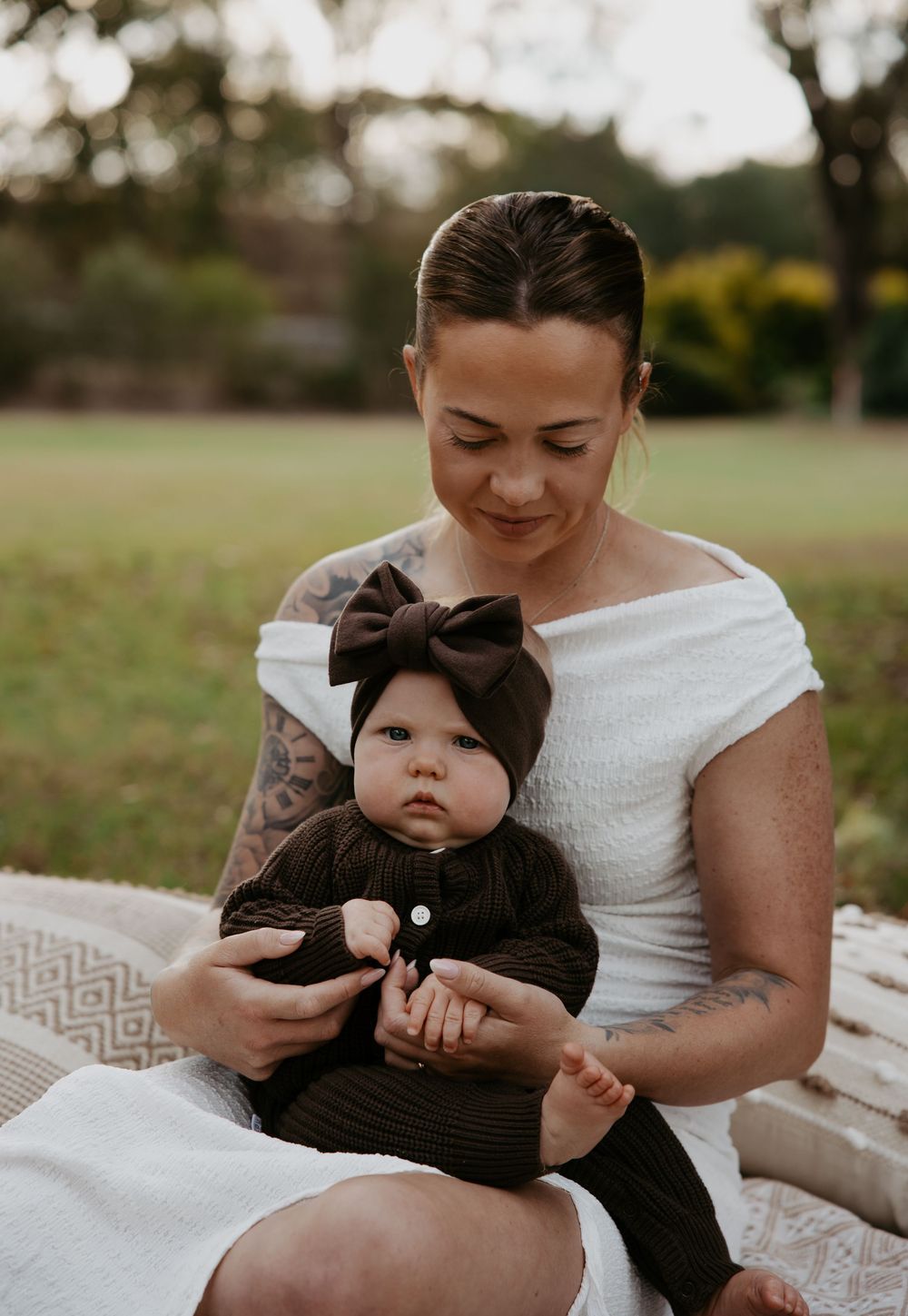 A tattooed mother holds her baby dressed in dark brown knit outfit with bow headband outdoors.