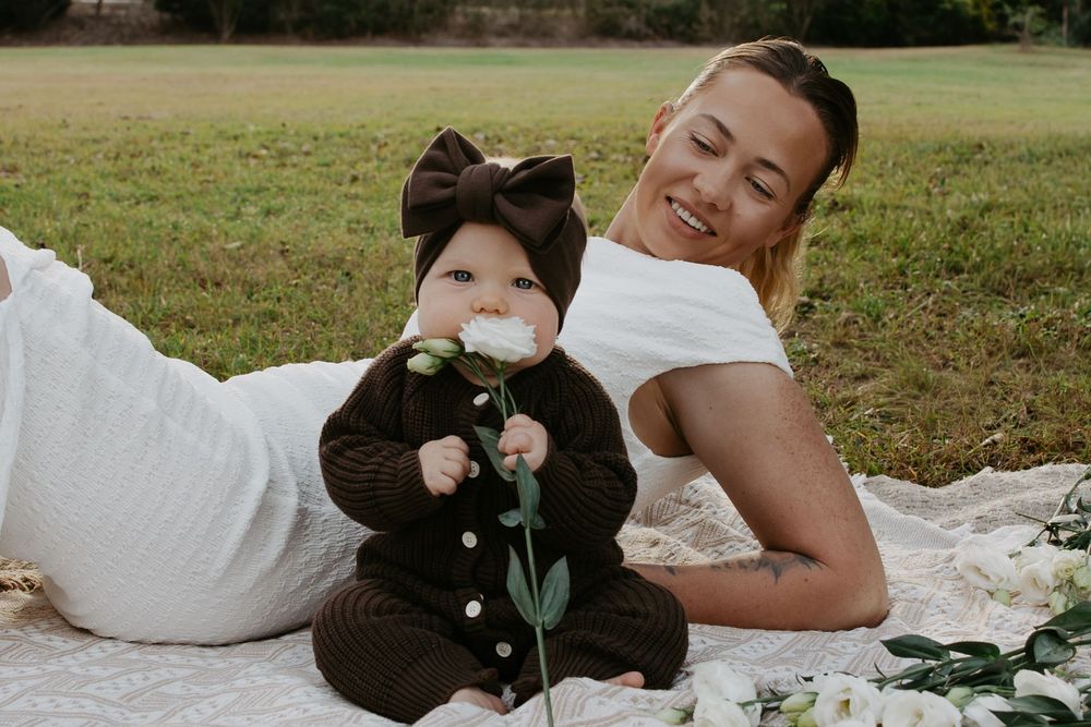 Smiling mother lying on white blanket outdoors with baby in brown knit outfit and bow headband holding a flower.