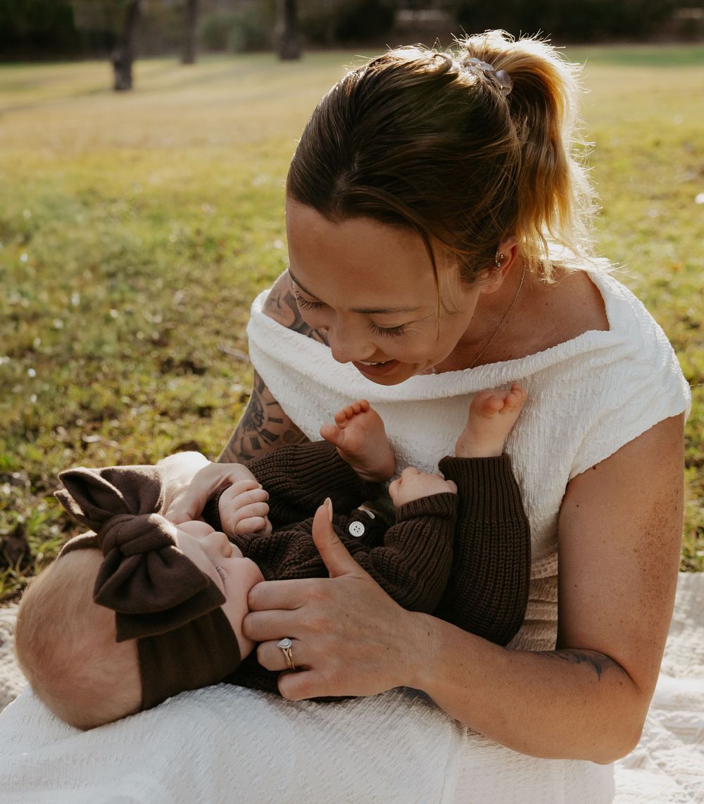 A mother tenderly breastfeeds her newborn baby outdoors on grass, wearing a white top and brown outfit.