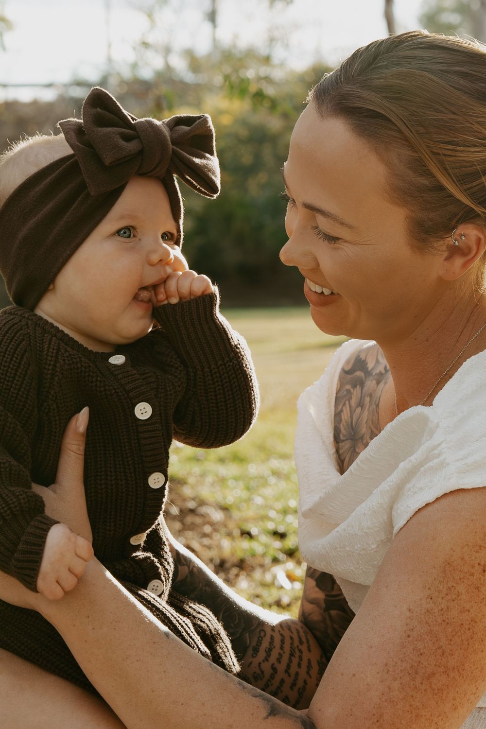 A smiling mother holds her baby girl wearing a black bow headband and knit outfit outdoors in warm sunlight.