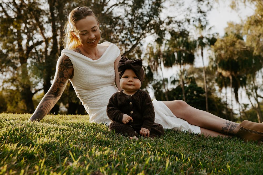 Tattooed mother sitting on grass smiling at her baby dressed in dark outfit with bonnet outdoors.