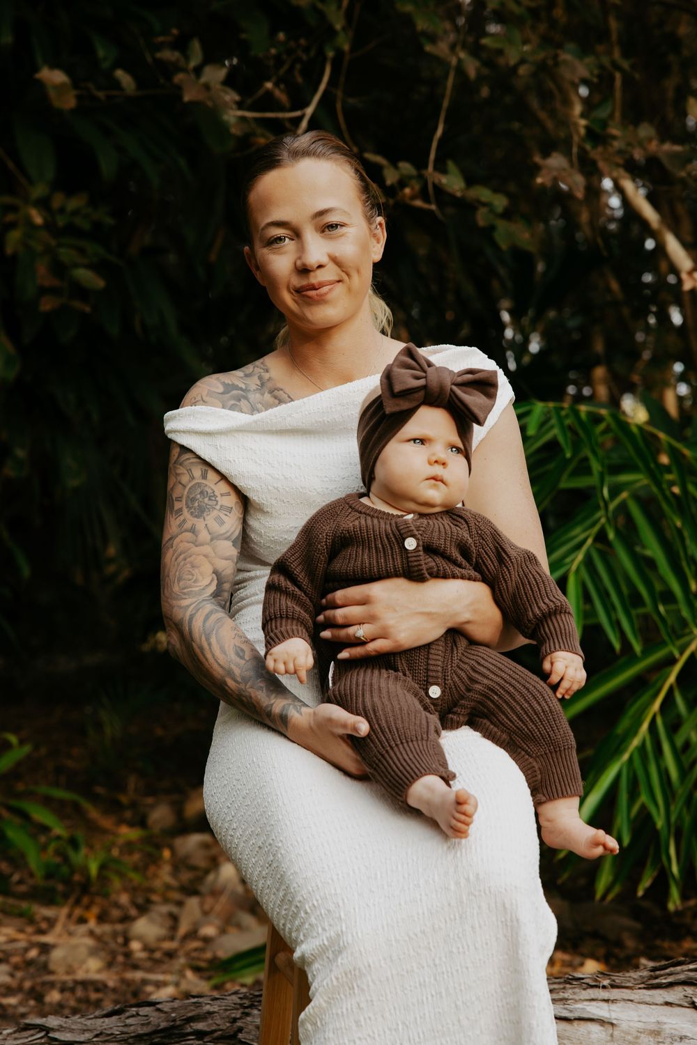 Tattooed mother in white dress holds baby in brown knit outfit with bow headband, seated outdoors.