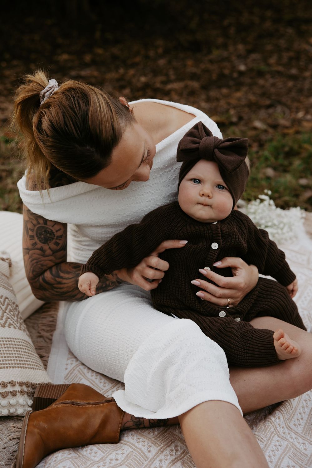 Tattooed mother in white dress holding her baby in a brown knit outfit with bow headband on a blanket outdoors.