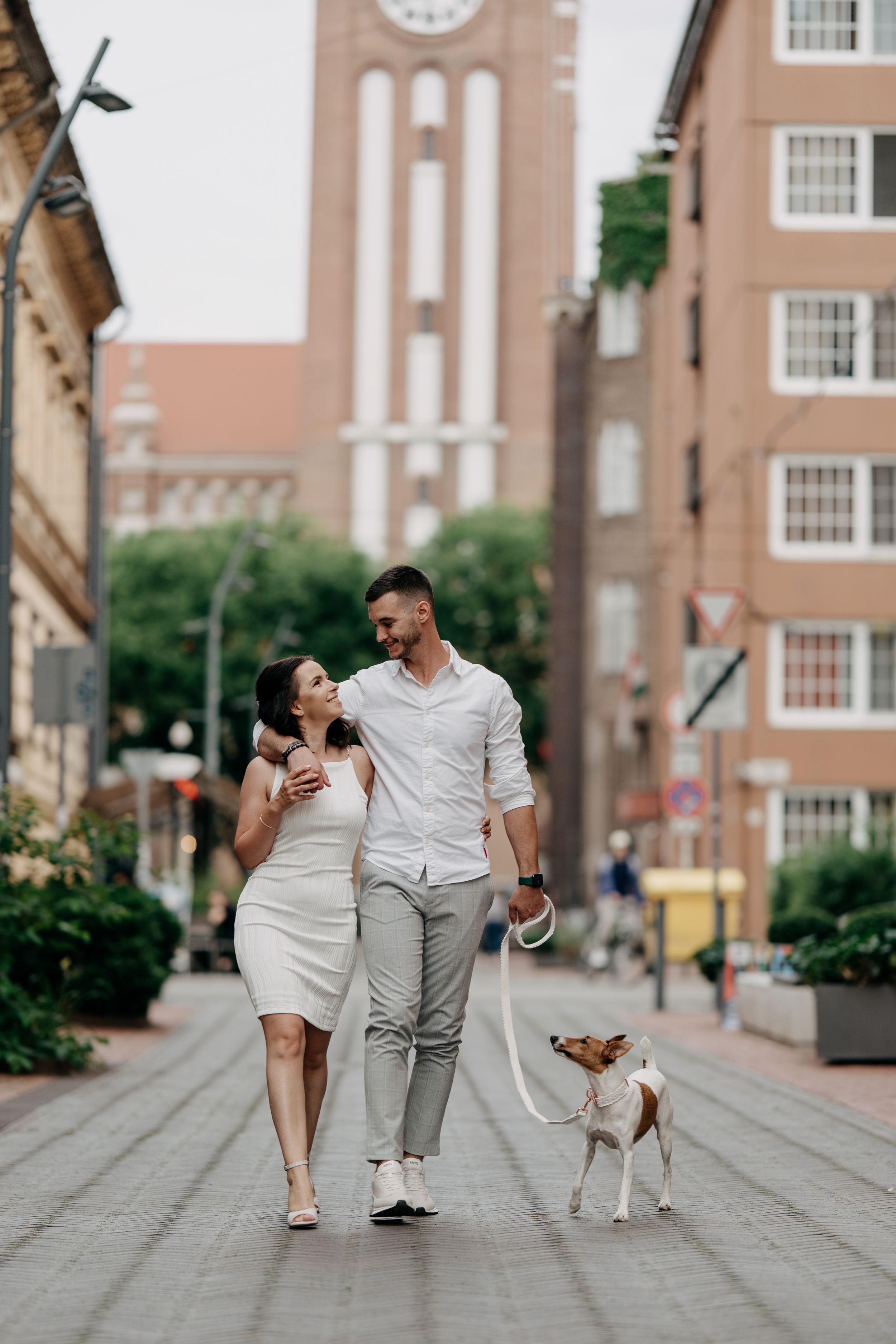 Couple walking dog on city street, woman in white dress, man in white shirt, brick buildings background.