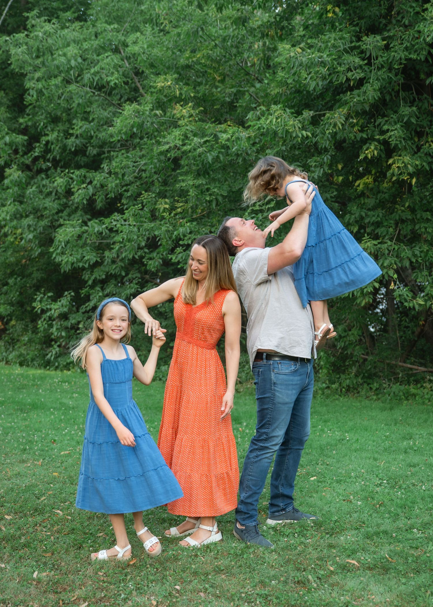 Family of four posing outdoors on green grass, two girls in blue dresses, mom in orange, dad lifting child.