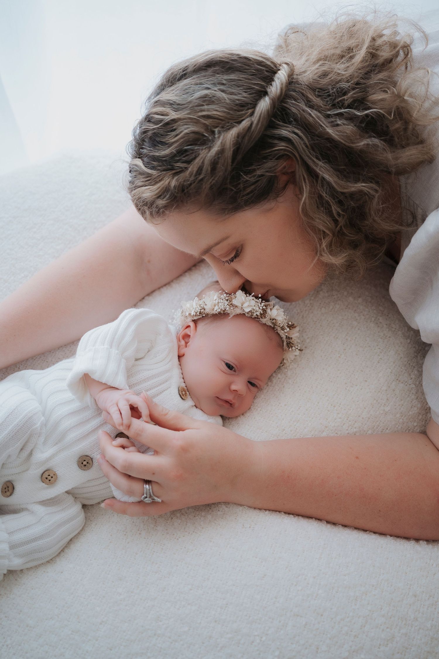 Mother tenderly nuzzling her newborn baby girl wearing a floral crown, both dressed in white, lying on a soft white surface.