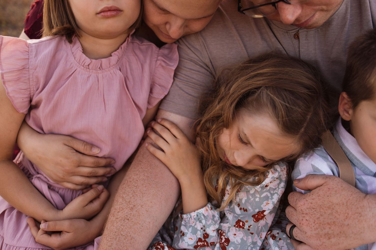 Family huddled together in warm embrace, parents cuddling two young girls in pink dresses outdoors.