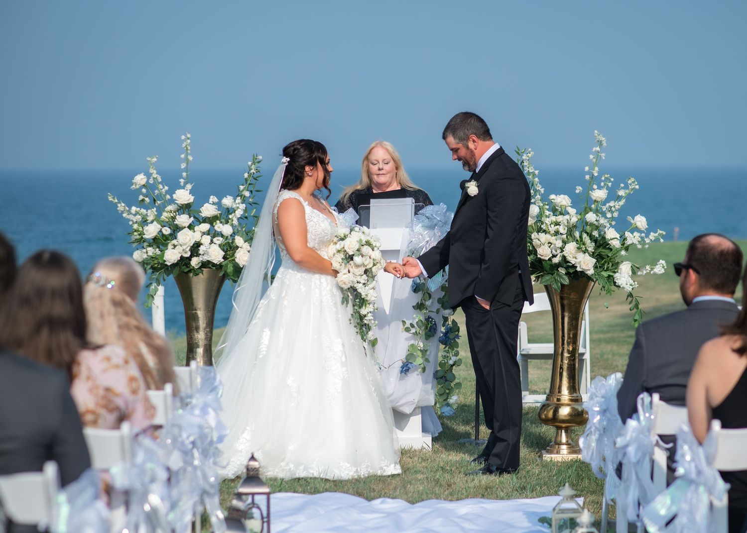 Bride and groom exchange vows at outdoor oceanfront wedding ceremony with white floral arrangements and seated guests.