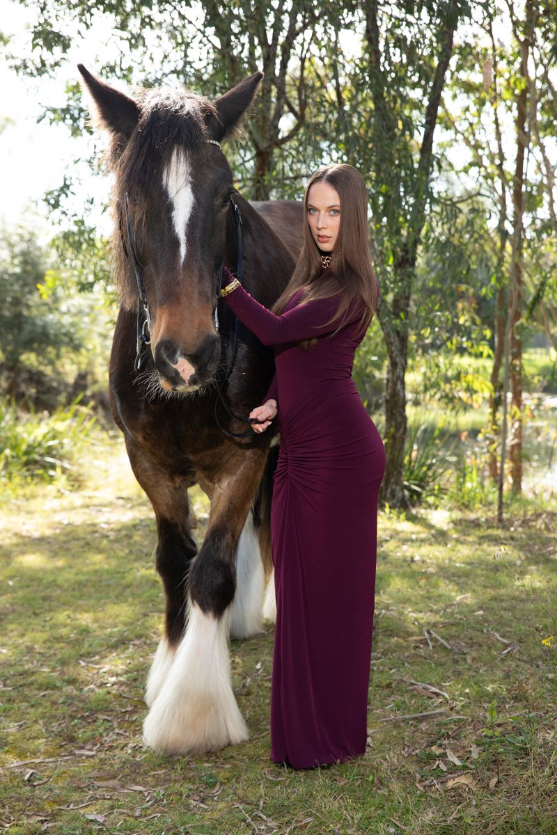 A woman in a burgundy gown stands beside a brown and white horse in a sunlit forest setting during a photoshoot in sydney