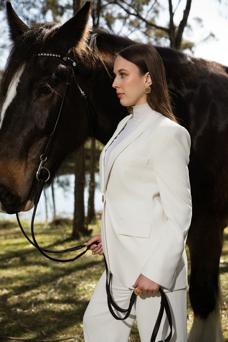 Woman in white suit holding reins beside a dark brown horse outdoors among trees during a photoshoot with a horse in sydney