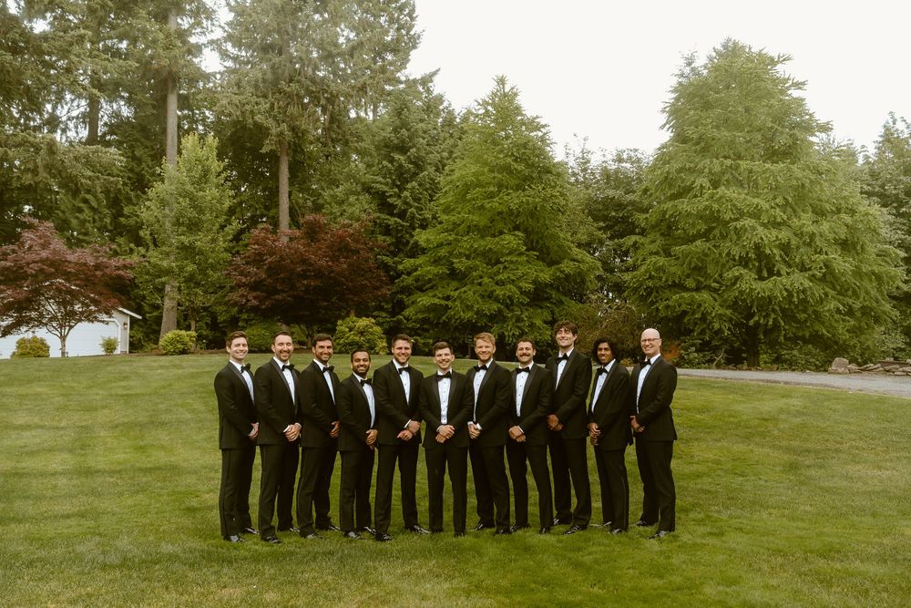 Group of groomsmen wearing matching black tuxedos standing on grass with evergreen trees behind.