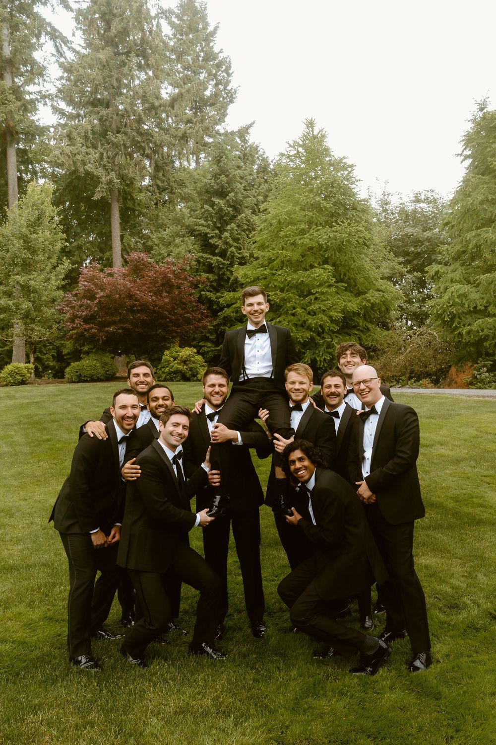 Groom lifted by groomsmen in black tuxedos on lush green lawn with tall evergreen trees backdrop.