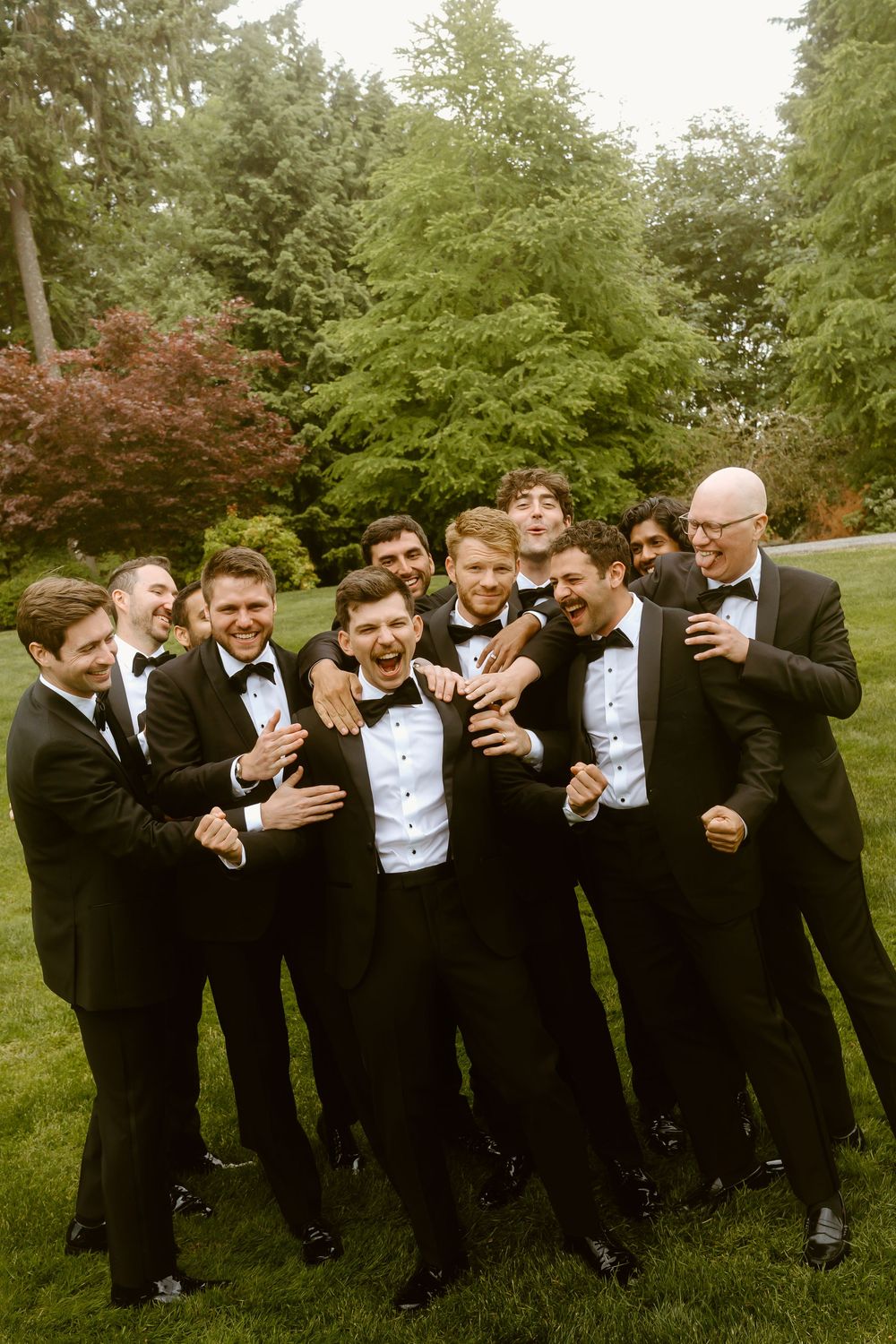 Groomsmen in black tuxedos laughing together outdoors on green lawn with lush trees in background at wedding.