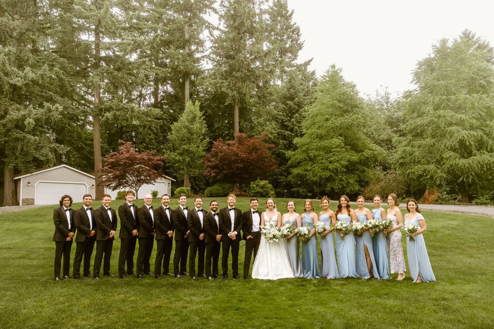 Wedding party poses outdoors, groomsmen in tuxedos and bridesmaids in blue dresses, with bride and groom centered on green lawn.