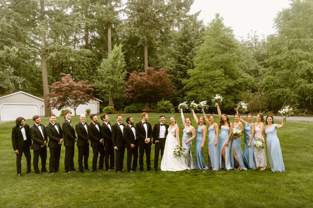 Wedding party poses outdoors, groomsmen in black tuxedos left, bridesmaids in blue dresses right, bride and groom centered.