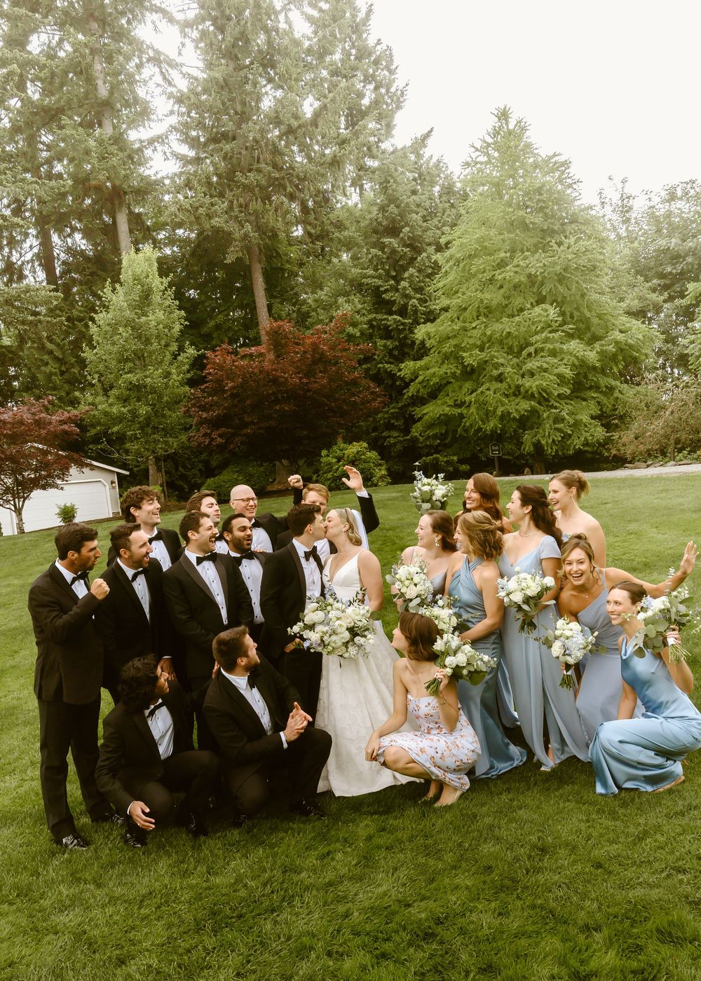 Wedding party in black tuxedos and blue dresses celebrate with bride on green lawn outdoors.