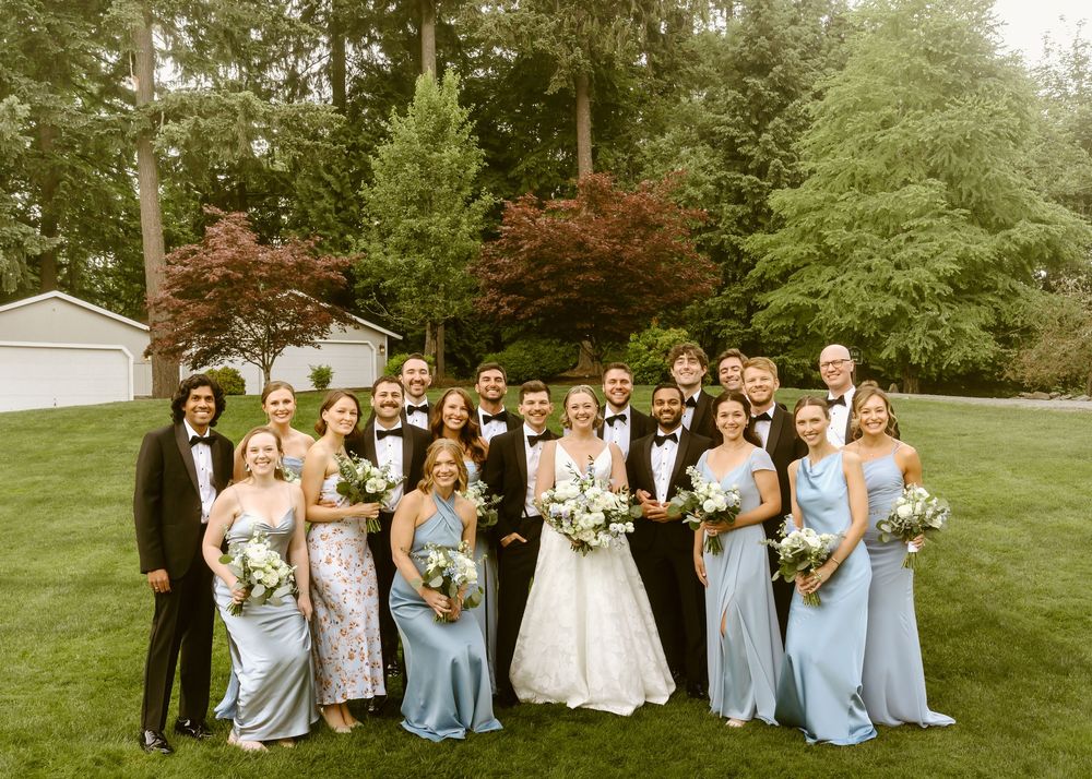 Wedding party posing outdoors in lush green setting, bridesmaids in blue gowns, groomsmen in black tuxedos, bride in white gown.