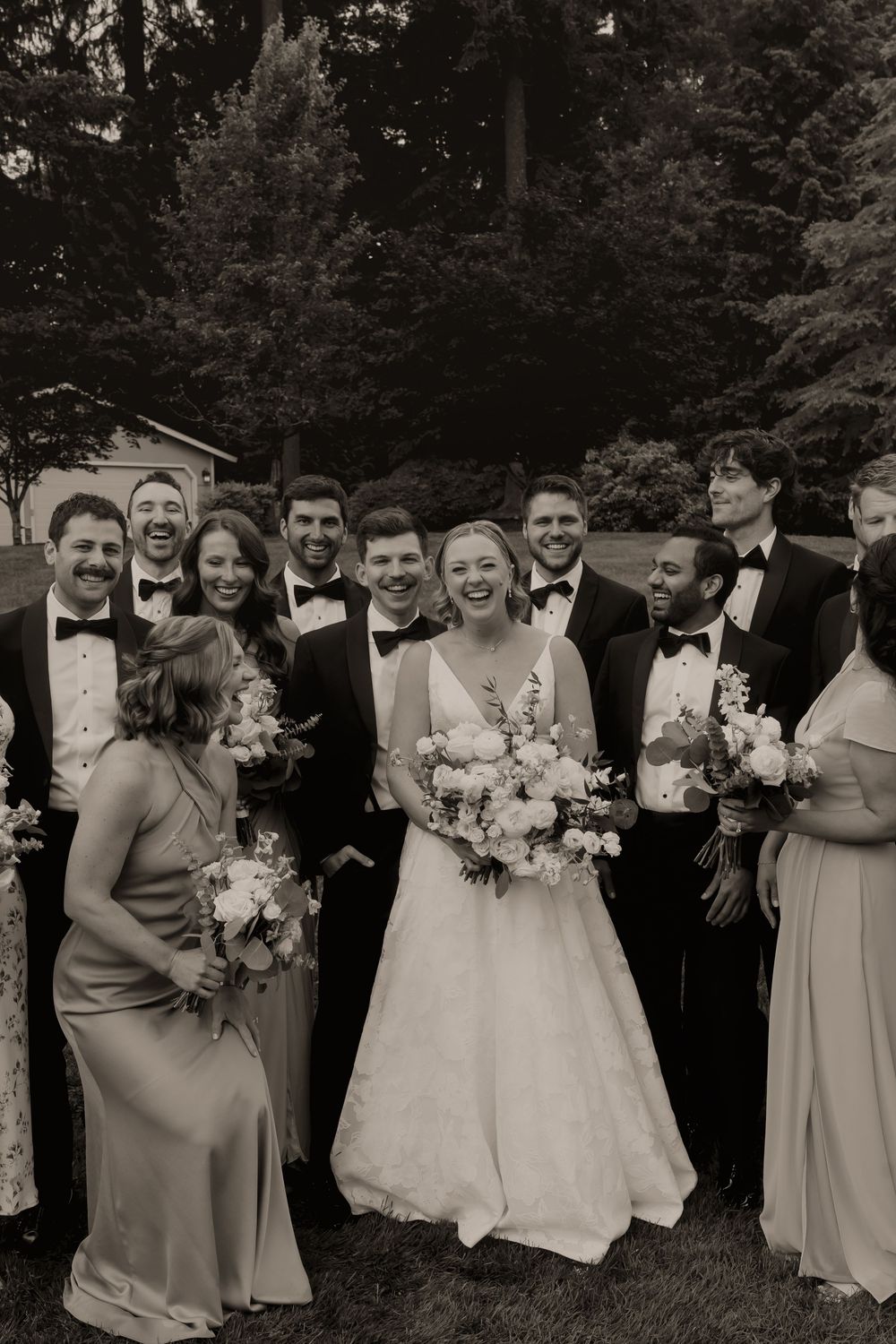 Black and white wedding party photo with bride holding bouquet surrounded by bridesmaids and groomsmen in tuxedos outdoors.