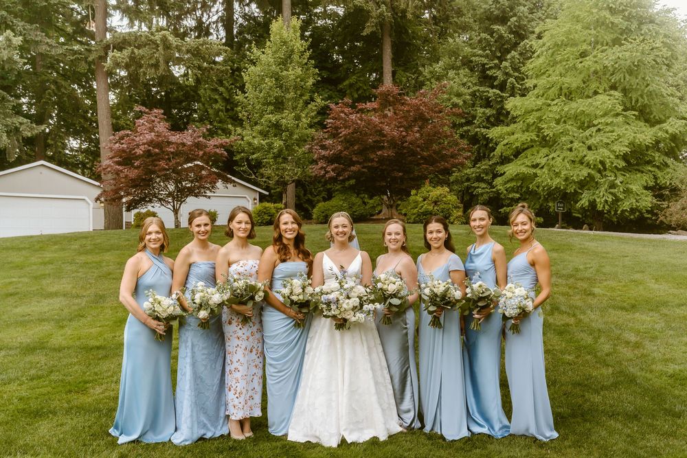 Bride in white gown poses with eight bridesmaids in light blue dresses holding bouquets on green lawn.