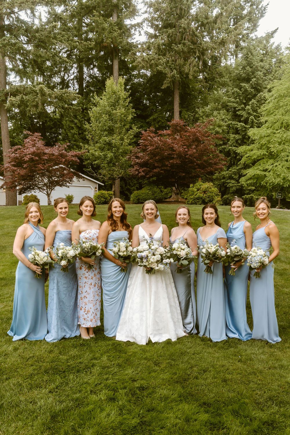 Bride in white gown poses with eight bridesmaids in blue dresses holding white bouquets on a green lawn.