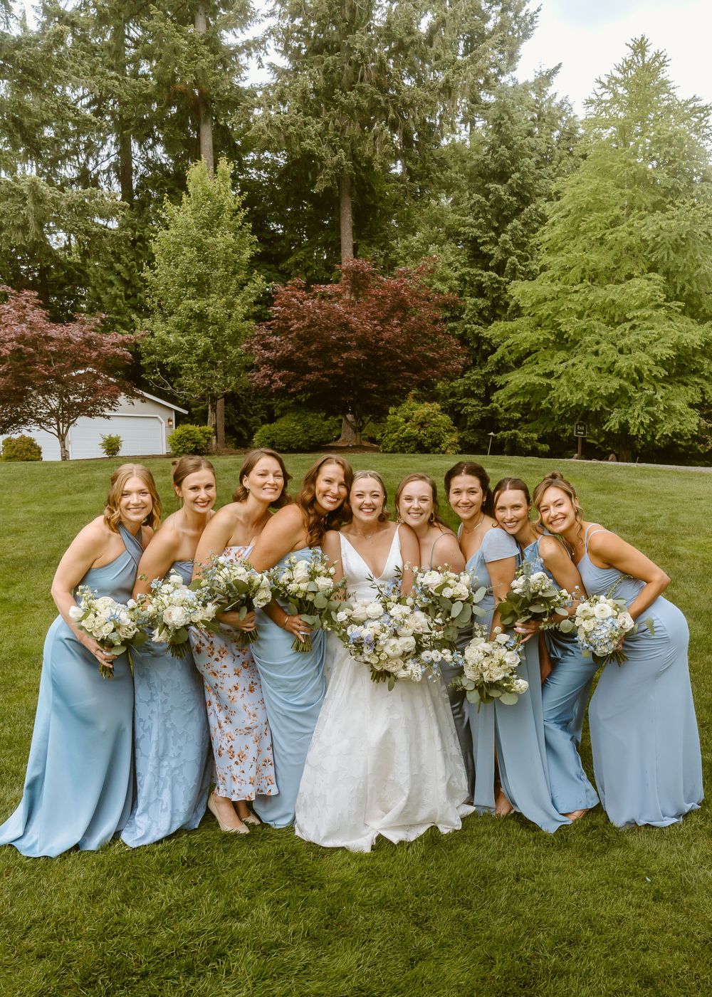 Bride in white gown poses with eight bridesmaids in blue dresses holding white bouquets on a green lawn.