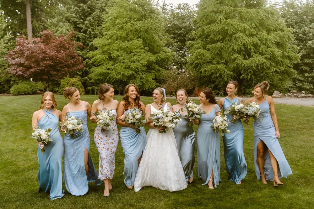 Bride and bridesmaids in blue satin dresses holding white bouquets, laughing together in a lush garden setting.