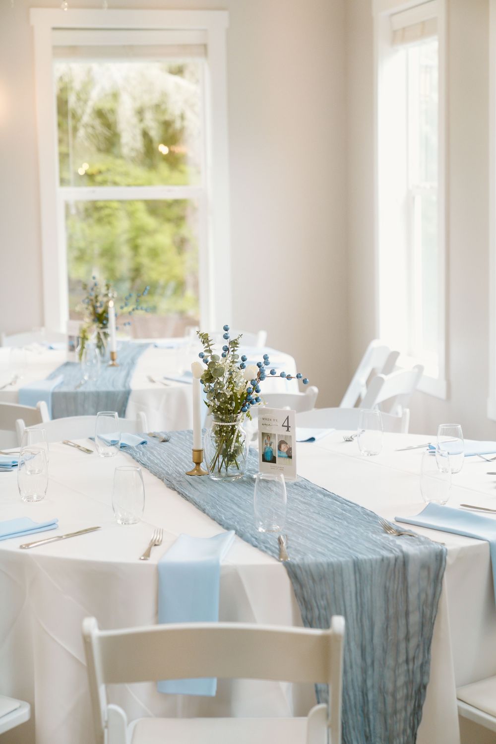 Elegant wedding reception tables with blue cheesecloth runners, floral centerpieces, and white chairs in bright venue.