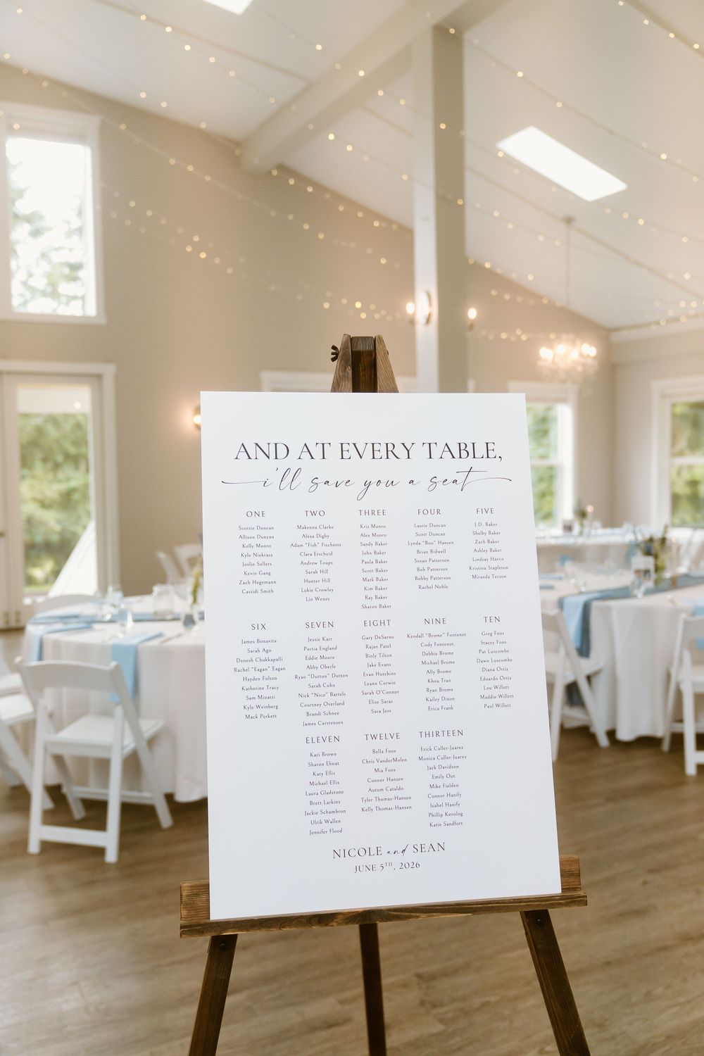 Wedding seating chart on easel reads 'And at every table, I'll save you a seat' in bright reception hall.