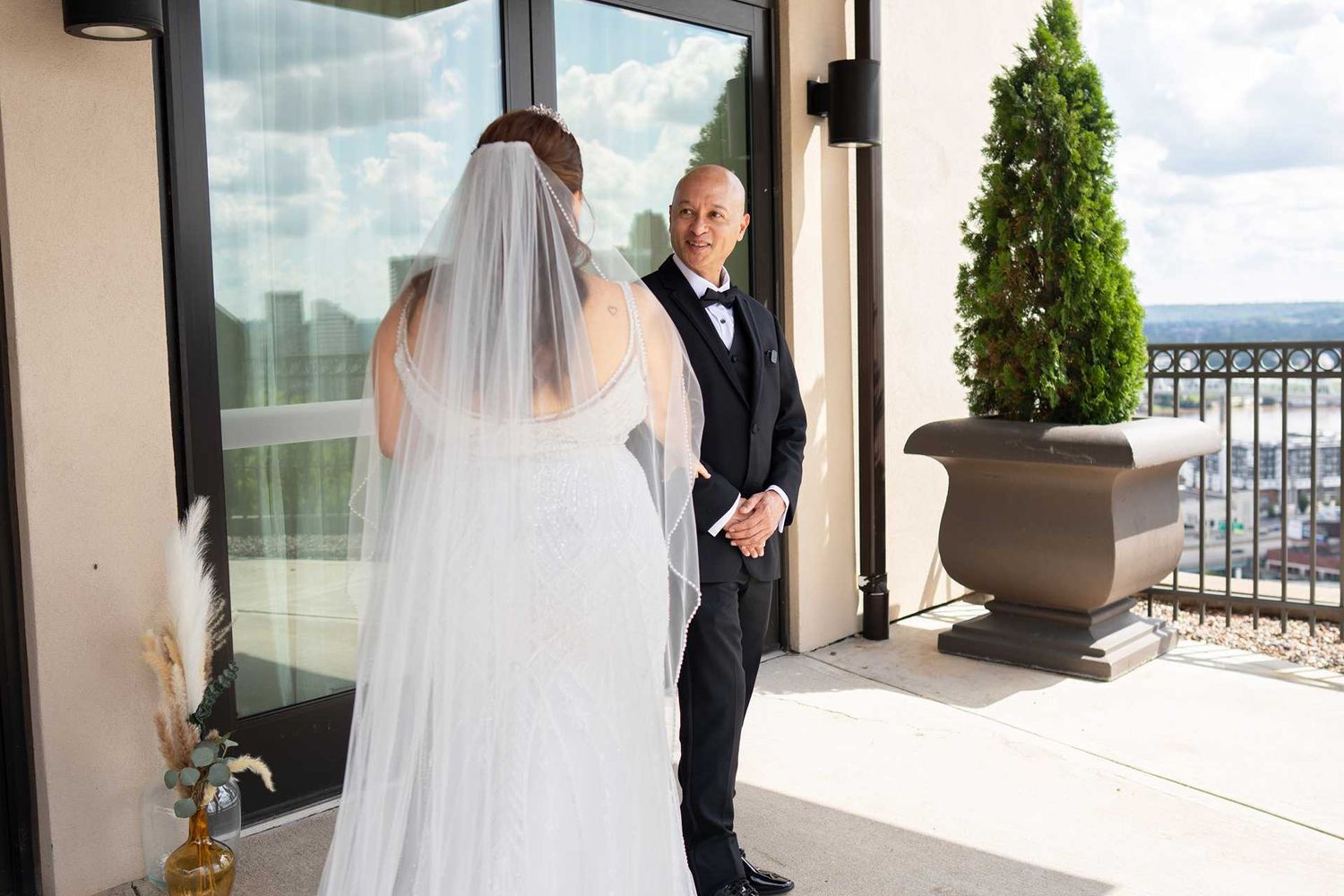 Bride in white gown and veil shares emotional first look moment with groom in black tuxedo on rooftop venue balcony.