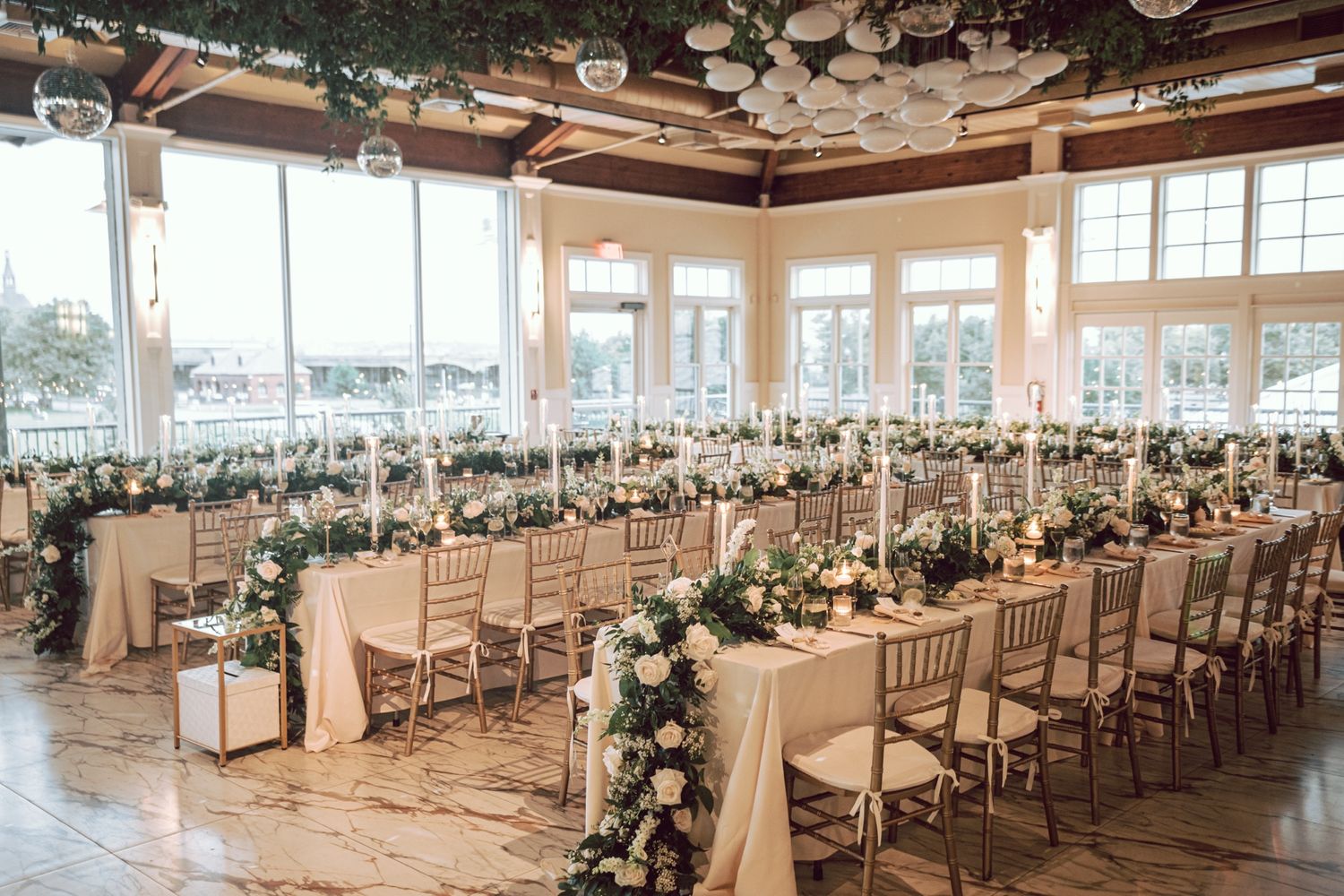 Elegant wedding reception hall with floral centerpieces, candles, gold chiavari chairs, and white tablecloths by large windows.